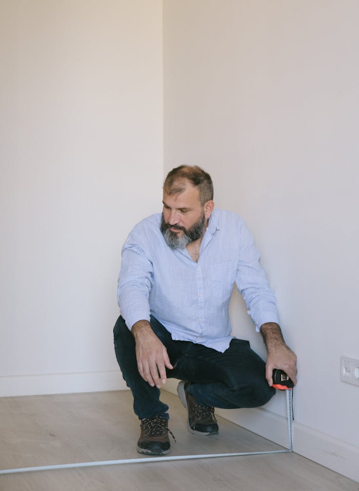 The Art of Drawing Readers In: Your attractive post title goes here A bearded man kneels on a wooden floor, measuring a room indoors.